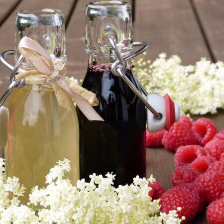 Lahodný domáci malinový sirup vo fľašiach s čerstvými malinami a kvetmi na drevenom stole.