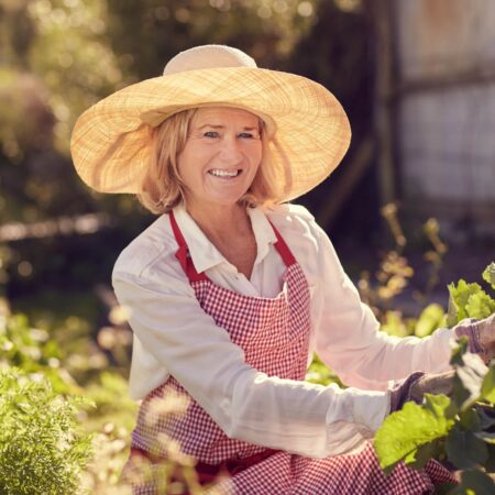 Záhradka a Alzheimerova choroba. Staršia žena s klobúkom a zásterou pracuje v záhrade, obklopená zelenými rastlinami.