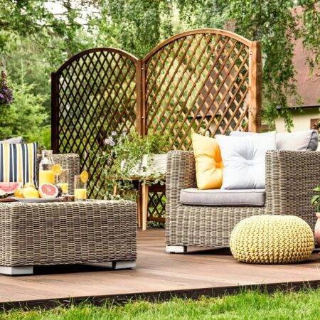 Drevená terasa s pohodlným ratanovým nábytkom, dekoratívnymi vankúšmi a osviežujúcimi nápojmi, obklopená zeleňou.