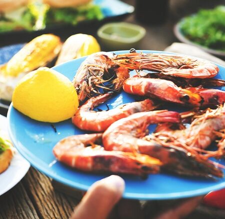 Strava v cudzine. Modrý tanier plný grilovaných kreviet a citrónov na stole s rôznymi jedlami v pozadí.