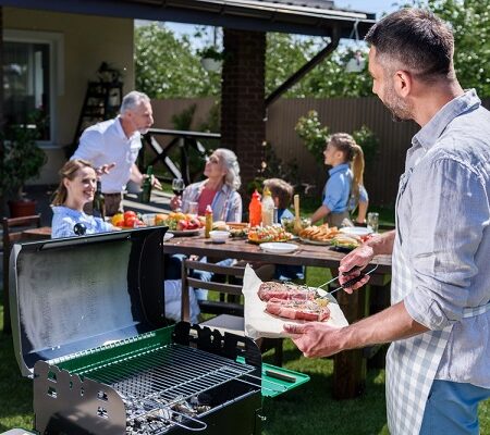 Rodina počas letnej záhradnej párty pri grilovaní, muž pri grile pripravuje jedlo, zatiaľ čo ostatní sa bavia pri stole.