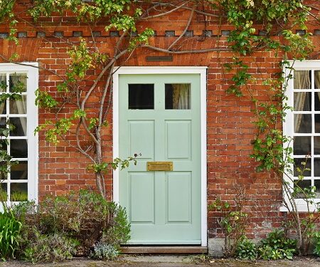 Tradičný červený tehlový dom s bielymi drevenými oknami a svetlo zelenými dverami obklopenými popínavými rastlinami.