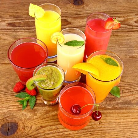 Farebné ovocné šťavy v pohároch na drevenom stole, obklopené čerstvým ovocím.