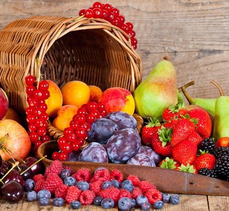 Je ovocie zlé? Colorful assortment of fresh fruits including apples, pears, strawberries, cherries, raspberries, blueberries, plums, and red currants spilling from a wicker basket on a rustic wooden table.