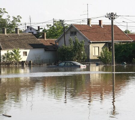 Škody po povodniach. Záplavená ulica s vodou dosahujúcou po strechy domov a auto zaplavené povodňovou vodou