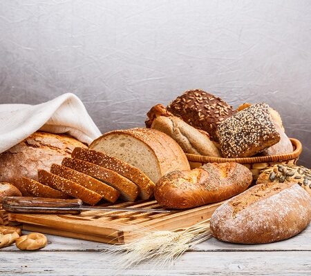 Chlieb a pečivo, Rôzne druhy čerstvého chleba a pekárenských výrobkov vrátane krájaného celozrnného mäsa, žemlí so semienkami a rustikálnych bochníkov na drevenom stole.