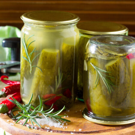 Kvasené uhorky v zaváraninových pohároch s rozmarínom, cesnakom a čili papričkami na drevenom podnose.