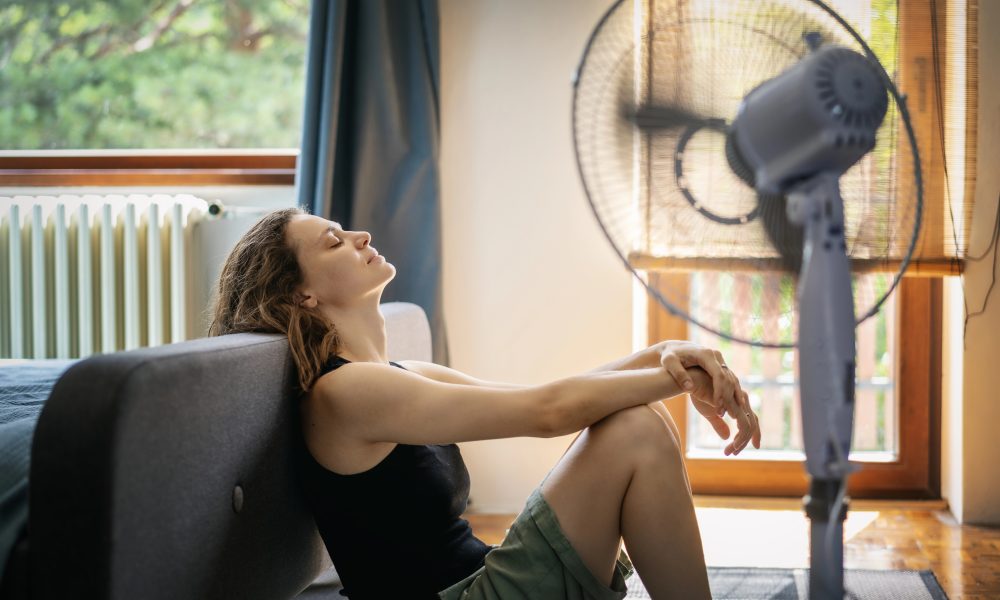 Ako schladiť vzduch v byte. Žena sa ochladzuje pri ventilátore počas letných horúčav v byte bez klimatizácie.