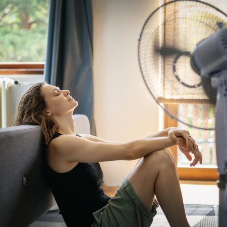 Ako schladiť vzduch v byte. Žena sa ochladzuje pri ventilátore počas letných horúčav v byte bez klimatizácie.