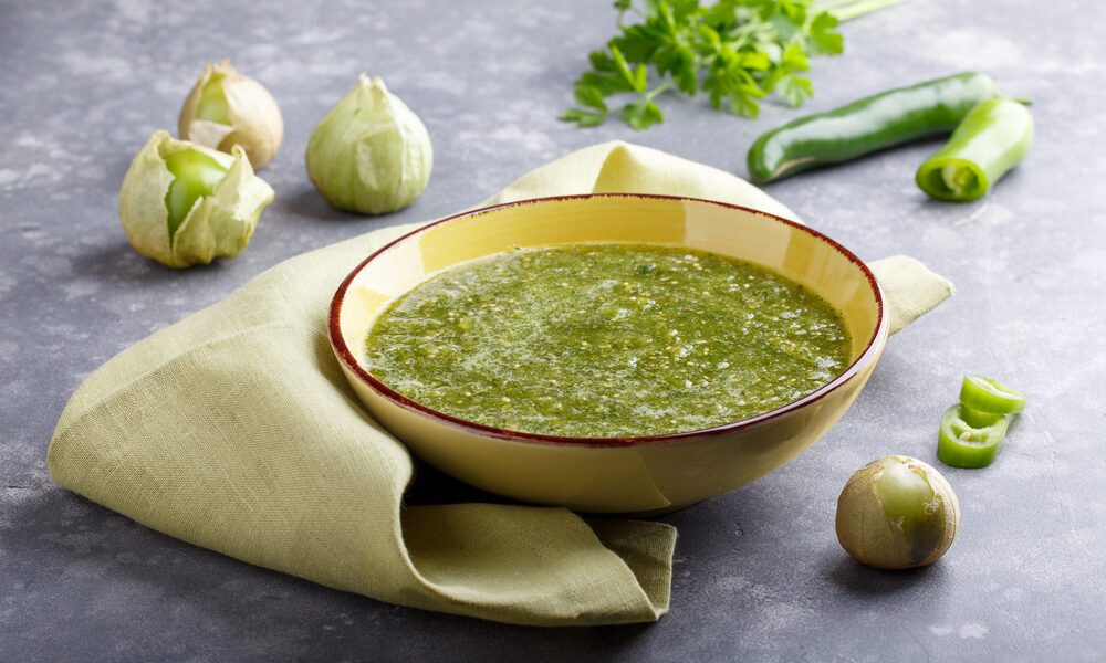 Zelená domáca salsa verde v žltej miske s tomatillos, zelenými papričkami a bylinkami v pozadí.