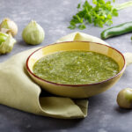 Zelená domáca salsa verde v žltej miske s tomatillos, zelenými papričkami a bylinkami v pozadí.